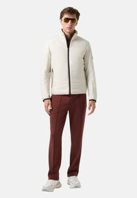 Lichtbeige gewatteerde jas met zwarte rits, gedragen over een donker shirt; gecombineerd met bordeauxrode broek en witte sneakers. Bruine zonnebril op model.