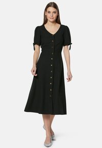 Schwarzes Midikleid mit V-Ausschnitt, kurzen Puffärmeln und durchgehender Knopfleiste. Es verfügt über goldene Knöpfe und einen fließenden Rock.