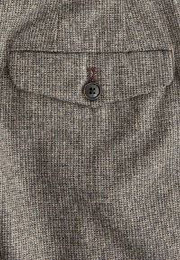 Gros plan sur la poche d'une veste en tweed marron et beige avec un rabat boutonné et une couture de boutonnière visible.