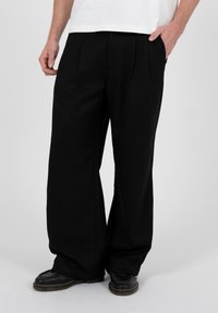 Person, die weite schwarze Hosen, schwarze Schuhe und ein weißes Hemd trägt und mit einer Hand in der Tasche vor einem einfarbigen Hintergrund steht.