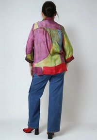 Mehrfarbige transparente Bluse mit einer Mischung aus rosa, grün und roten Farbtönen. Lockere Passform, lange Ärmel und abgerundeter Saum. Kombiniert mit geraden blauen Jeans und roten Heels.