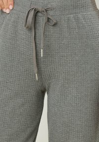 Close-up van grijze ribgebreide broek met een getrokken koord en een deel van een hand die op de tailleband rust.