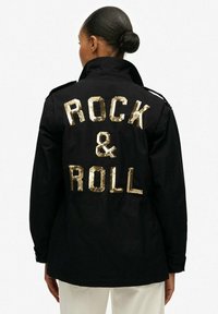 Chaqueta negra con letras de lentejuelas doradas en la parte trasera que dicen "ROCK & ROLL". Presenta un cuello clásico y epaulettes en los hombros.