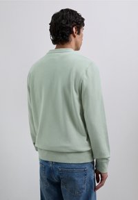 Sudadera de cuello redondo verde claro con mangas largas, hecha de una tela suave, con puños y dobladillo de canalé, llevada sobre unos vaqueros de mezclilla azules.