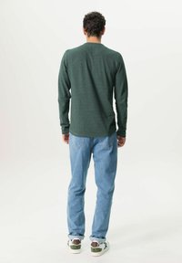 Chemise verte à manches longues avec un col rond, associée à un jean bleu clair et des baskets avec des accents bruns et blancs, vue de l'arrière.