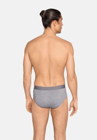 Homme aux cheveux longs attachés en chignon, portant un slip gris avec une ceinture de marque, debout de dos sur un fond uni.