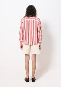 Gestreiftes Baumwollhemd in Rot und Creme, mit langen Ärmeln und lockerer Passform. Kombiniert mit off-weißem Jeansshorts mit fransigen Kanten.