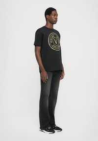 Tricou din bumbac negru, cu un logo circular mare de culoare aurie pe care este inscripționat textul "VERSACE JEANS COUTURE". Blugi gri și adidași negri completează ținuta.