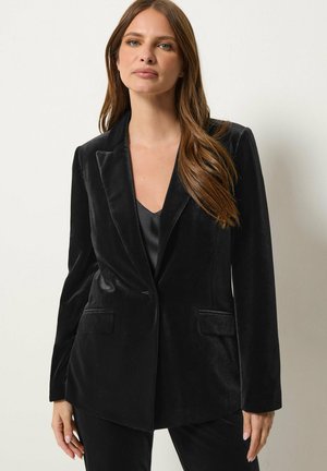 Femme aux longs cheveux bruns porte un blazer en velours noir sur un haut noir, debout devant un fond clair uni.