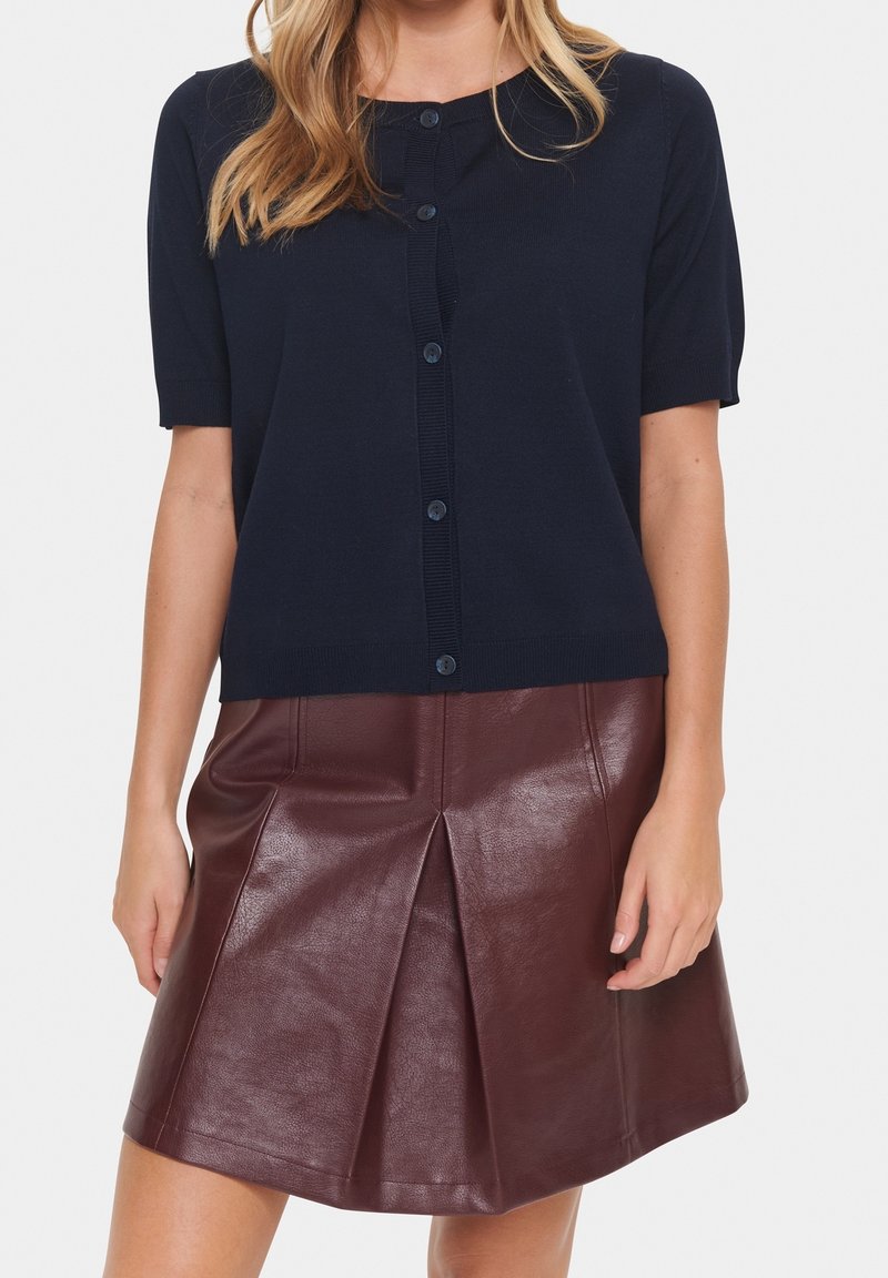 Cardigan bleu marine à manches courtes avec boutons et texture côtelée, associé à une jupe en cuir bordeaux à plis avec une finition lisse.