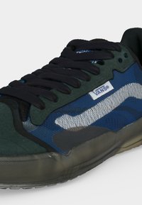 Zbliżenie na ciemnozieloną i niebieską tenisówkę Vans z czarnymi sznurowadłami i przezroczystą podeszwą, z logo Vans na bocznej części buta.