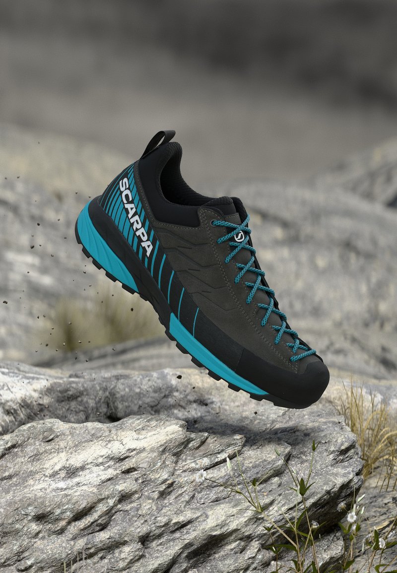 Μαύρο και μπλε πεζοπορικό παπούτσι SCARPA κρεμασμένο πάνω από βραχώδες έδαφος με μικρά φυτά, τονίζοντας τα χαρακτηριστικά σχεδίασης για υπαίθριες και ανθεκτικές χρήσεις.