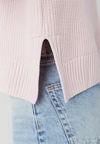 Pull rose clair en maille avec ourlet côtelé et fentes latérales, associé à un denim bleu, mettant en valeur un motif de tissu texturé.