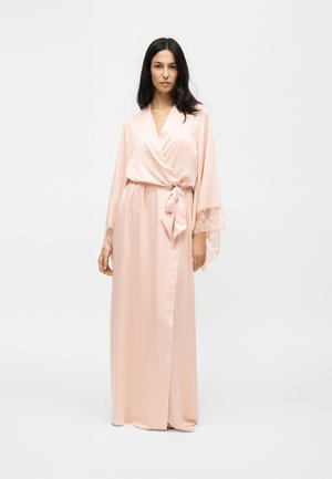 Femme aux longs cheveux foncés portant une robe portefeuille en satin rose clair avec des manches longues bordées de dentelle, nouée à la taille.
