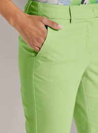 Hand mit heller Hautfarbe, die in der vorderen Tasche einer limettengrünen Hose ruht, vor einem einfarbigen Hintergrund.
