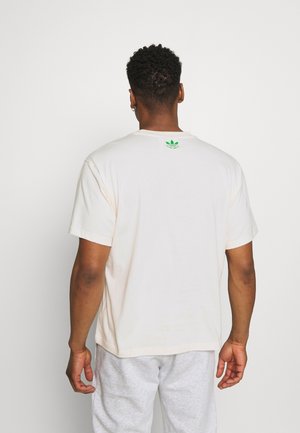 Mand med kort krøllet hår iført en hvid T-shirt med et lille grønt Adidas-logo på øvre ryg, kombineret med lysegrå joggingbukser.