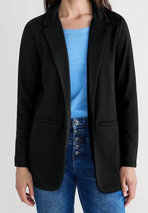 Kvinde iført en sort blazer over en blå top med højtaljede blå jeans med fire synlige knapper.
