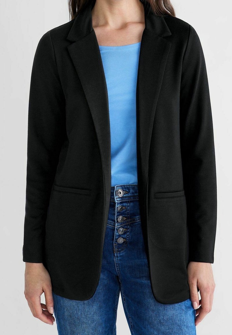 Femme portant un blazer noir sur un top bleu avec un jean bleu taille haute comportant quatre boutons visibles.