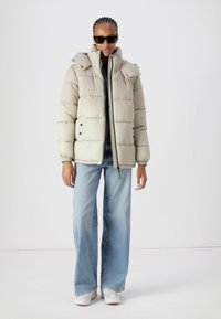 Pufferjakke i lys beige med høy krage, trykknapper og sidelommer. Kombinert med blå vide jeans og hvite joggesko.