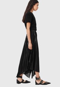 Musta pörröinen lyhythihainen paita yhdistetty mustaan, laskostettuun midihameeseen, jossa on korkea-matala helma ja kiiltävä pinta, täydentäen kokonaisuuden mustilla, solkikoristeisilla matalilla kengillä.