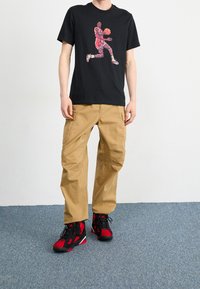 Camiseta negra con un gráfico de un jugador de baloncesto multicolor, pantalones cargo caqui y zapatillas deportivas rojas y negras con suelas texturizadas.