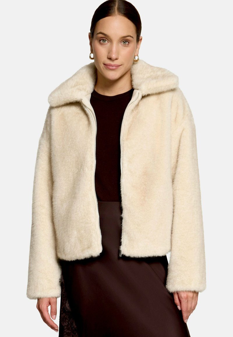 Femme portant une veste beige duveteuse zippée sur un top noir et une jupe marron foncé, debout devant un fond clair uni.