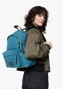 Zaino a fantasia teal con design a griglia, dotato di due tasche con zip, spallacci regolabili e manico superiore, indossato sopra una giacca oliva.