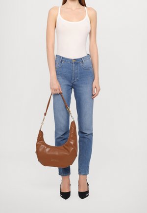 Femme portant un haut blanc sans manches, un jean bleu et des talons noirs, tenant un grand sac à main en cuir brun par sa sangle.