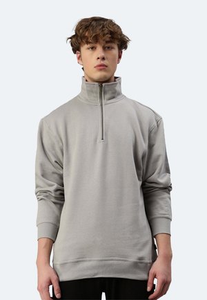 Giovane uomo con capelli ricci che indossa un maglione grigio chiaro con cerniera frontale e collo alto, in piedi davanti a uno sfondo semplice.