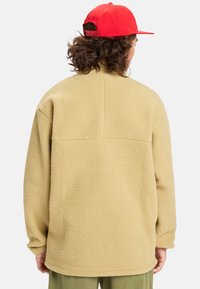 Beige fleece jacket with a textured surface, straight cut, and no visible fastenings, worn with a red cap in the background.