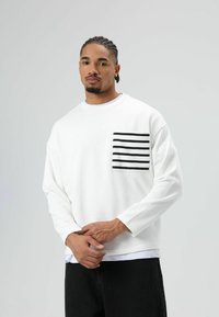 Witte trui met lange mouwen, voorzien van een zwart-wit gestreepte zak. Gestyled met donkere broek en een ronde hals.