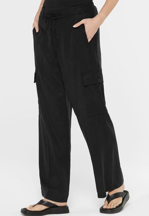 Person wearing loose black cargo pants with side pockets and black platform flip-flops, hands in pockets, against a white background.