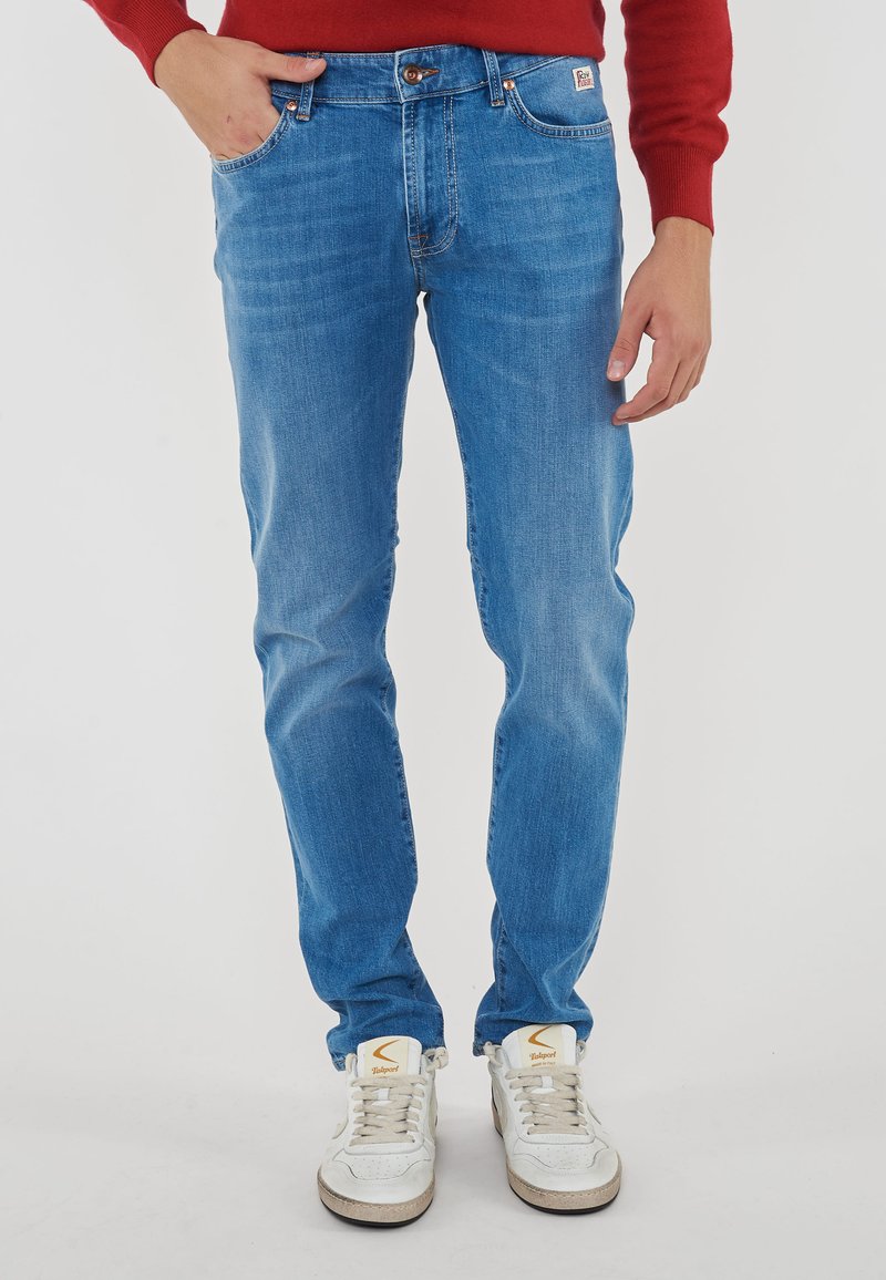 Persona che indossa jeans di denim blu, sneakers bianche con logo arancione e una maglietta rossa a maniche lunghe, in piedi contro uno sfondo semplice.
