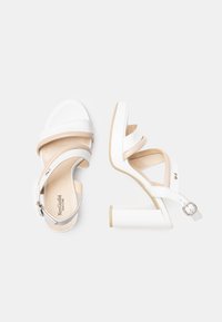 Sandales blanches à talons avec une finition lisse, une bride de cheville, et un talon block large, présentant deux larges brides sur le pied et des accents neutres.