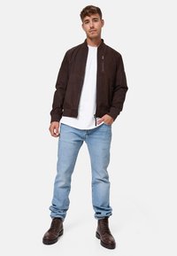 Braune Wildleder Bomberjacke mit gerippten Bündchen, Frontreißverschluss und einer Brusttasche, kombiniert mit hellblauen Jeans und braunen Schnürstiefeln.