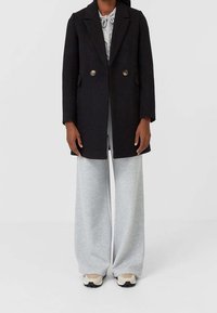 Zwarte wollen jas met een gestructureerd ontwerp, dubbele rij knopen en zijzakken, gedragen over grijze sweatpants met brede pijpen en sneakers.