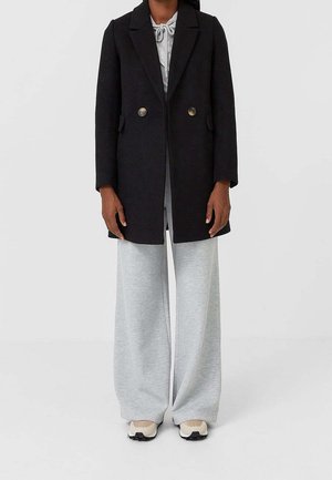 Black wool coat with a structured design, double-breasted buttons, and side pockets, worn over grey wide-leg sweatpants and sneakers.