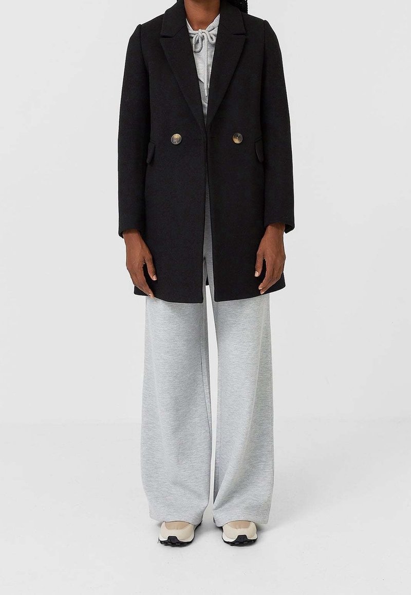 Zwarte wollen jas met een gestructureerd ontwerp, dubbele rij knopen en zijzakken, gedragen over grijze sweatpants met brede pijpen en sneakers.