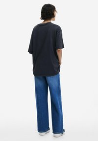 Übergroßes schwarzes T-Shirt mit kurzen Ärmeln, kombiniert mit lockeren blauen Jeans, von hinten vor einem weißen Hintergrund gezeigt.