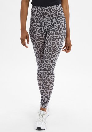 Legginsy z wysokim stanem w szarym wzorze w panterkę, z gładką fakturą i kontrastującymi czarnymi plamkami, w zestawieniu z białymi butami sportowymi.