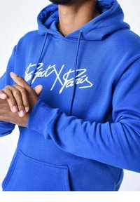 Uomo che indossa una felpa blu brillante con testo bianco, le mani congiunte davanti a sé e una barba visibile, su uno sfondo bianco.