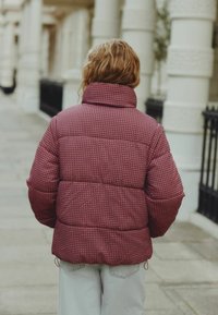 Bordeaux-farvet polstret jakke med høj krave og struktureret stof, sammen med lysegrå bukser, set fra ryggen i en urban setting.
