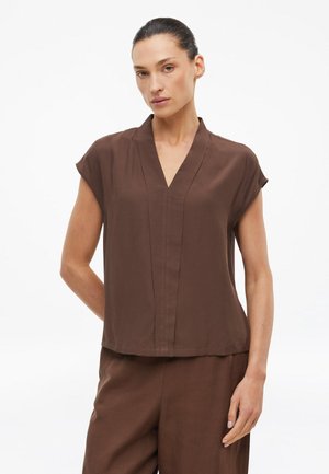 Vrouw met donker haar in een lage knot, gekleed in een bruine korte mouw V-hals top en bijpassende broek tegen een witte achtergrond.