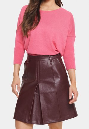 Frau trägt einen rosa Pullover mit langen Ärmeln, der in einen hoch taillierten, dunkelbraunen Lederrock in A-Linie mit vorderen Falten gesteckt ist.