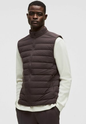 Donkerbruine puffergilet met een hoge kraag, met horizontaal gestikte secties en een volledige ritssluiting aan de voorkant, gedragen over een lichtgekleurde langemouwen T-shirt.