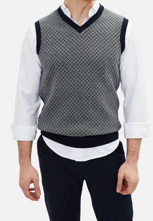 Mężczyzna ubrany w szary, wzorzysty bezrękawnik swetra z dekoltem w serek, na biały długi rękaw z podwiniętymi rękawami i ciemne spodnie.