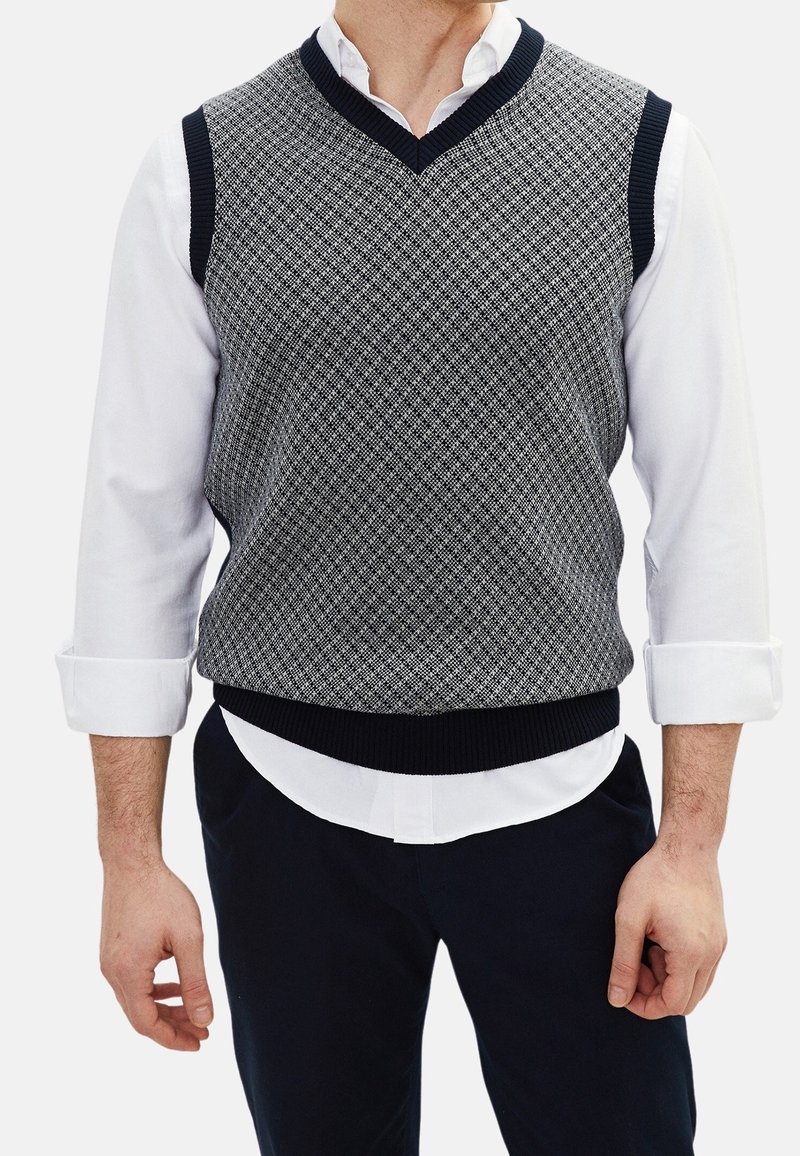 Homme portant un gilet sans manches gris à motif avec col en V par-dessus une chemise blanche à manches longues retroussées et un pantalon foncé.