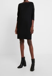 Robe noire à coupe ample, manches trois-quarts et encolure ronde. Caractérisée par des accents texturés subtils sur les épaules. Portée avec des bottines.