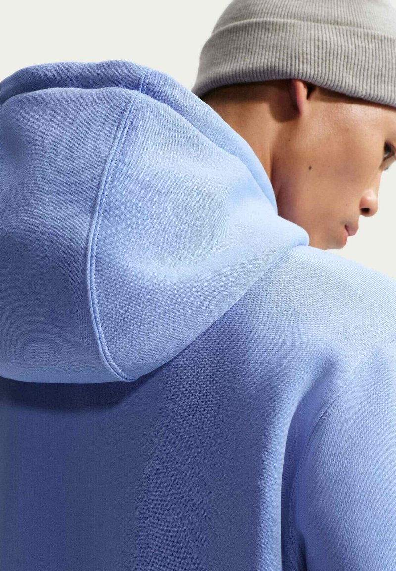 Sudadera con capucha azul claro con un acabado texturizado y capucha con cordón, mostrando un primer plano de los detalles de costura y un gorro gris al fondo.