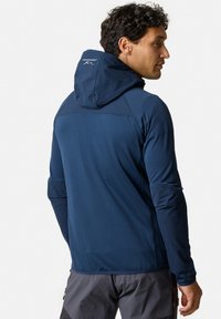 Marineblauwe sportjacket van een gestructureerde stof, met een aansluitende capuchon en contrasterend schouderpaneel. Voorzien van een logo op de achterkant.